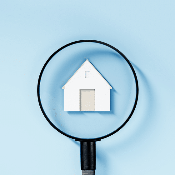 Wit huisje onder een vergrootglas op een lichtblauwe achtergrond, symbool voor het zoeken naar een woning