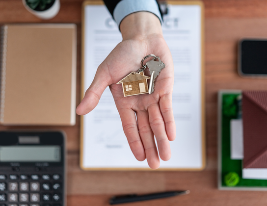 Hand met woningsleutel en houten huisje boven een contract voor woningbeheer in Voorschoten
